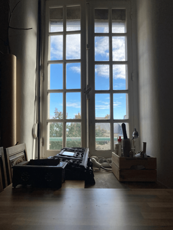Une vue intérieure d'une pièce avec une fenêtre ouverte, montrant un ciel bleu et des arbres à l'extérieur. Sur la table, il y a des objets divers, y compris un décor en bois et des ustensiles.