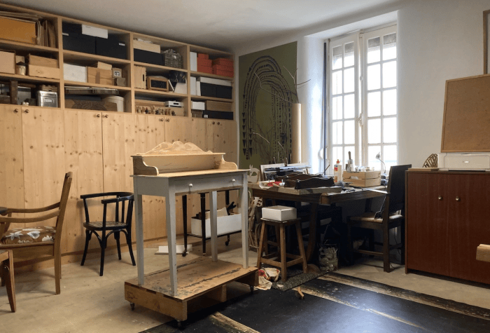 Un atelier de travail avec des meubles variés, incluant une table au centre et des étagères en bois remplis de fournitures, des sièges et un sol en bois.